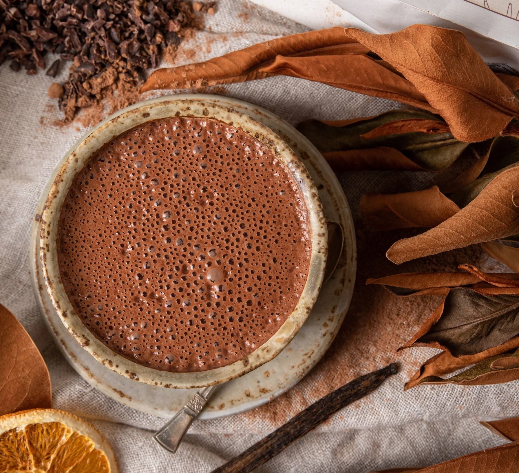 Cup of Mamamuti Cacao resting on table cloth alongside autumn leaves, orange and Cacao nibs